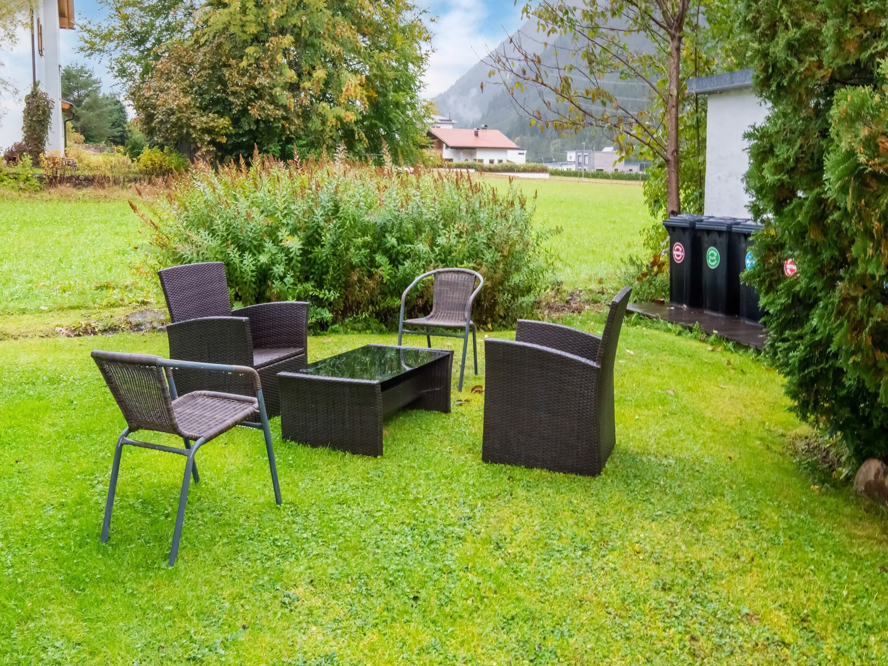Un área de estar al aire libre con mobiliario de mimbre, una mesa de vidrio y un pequeño jardín con arbustos y un árbol.
