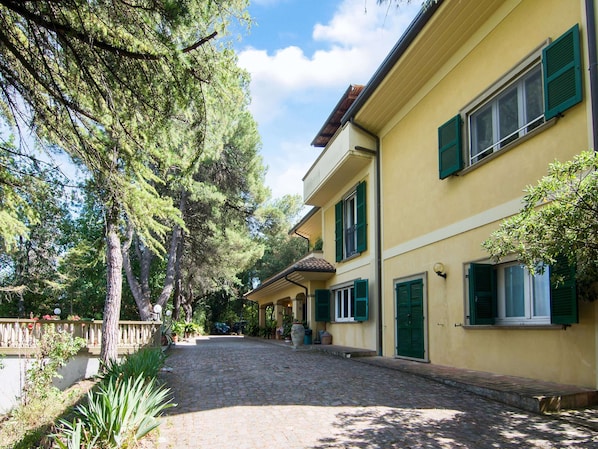 A yellow building with green shutters, a cobblestone driveway, and surrounding trees.