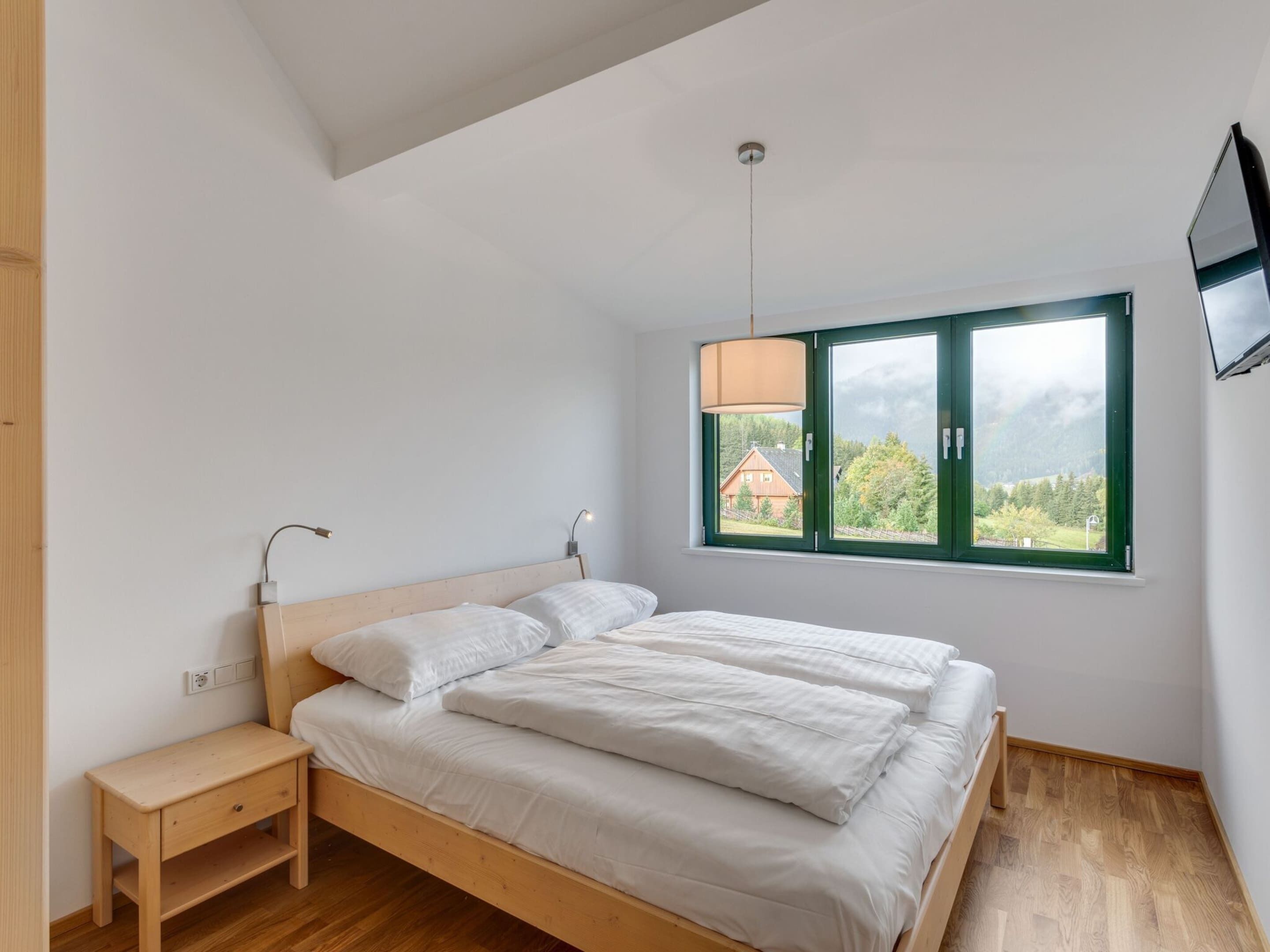 Une chambre à coucher avec un lit en bois, une table de chevet, une fenêtre donnant sur une maison et des arbres, et un luminaire au plafond.