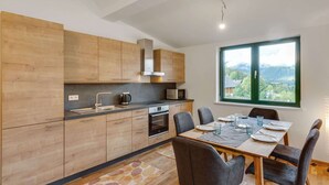 A modern kitchen with wooden cabinets, a dining table set for four, and a large window offering a view of a forested landscape. - Apartment in Hohentauern Near Ski Lift (Hohentauern)