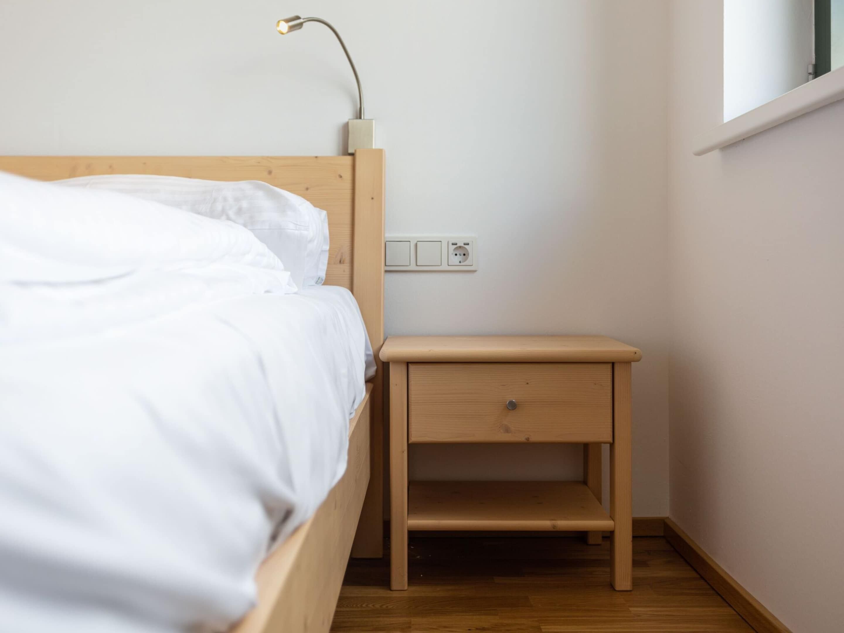 A bedroom with a wooden bed, a nightstand with a drawer, a lamp, and a power outlet on the wall.
