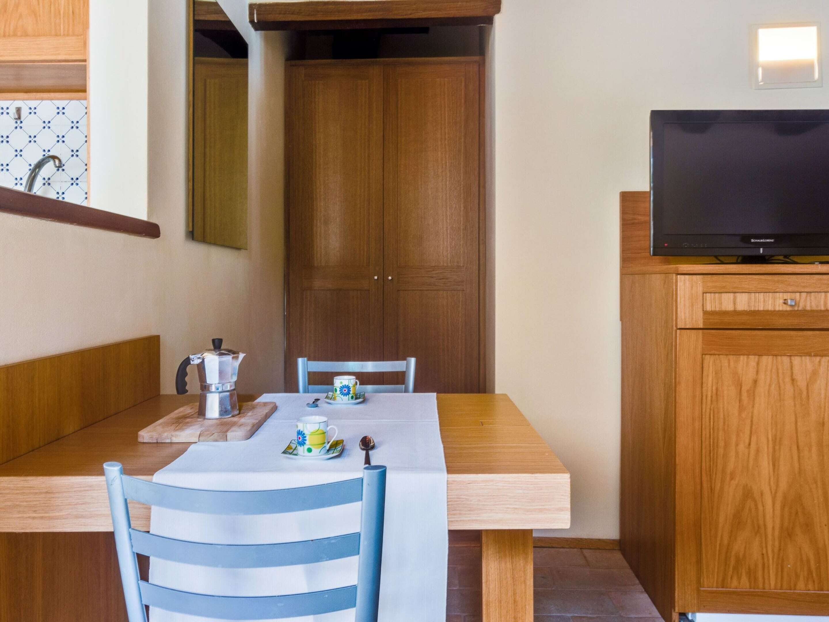 Un pequeño comedor con una mesa de madera, dos sillas, una cafetera y un televisor sobre un mueble de madera.