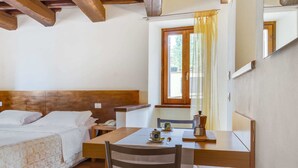 Ein Zimmer mit Bett, dessen Kopfteil aus Holz ist, ein kleiner Tisch mit Kaffeemaschine, ein Sessel und ein Fenster mit Vorhängen.