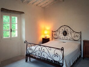 Un dormitorio con estructura metálica para la cama, una mesita de noche con lámpara y ventana con vista a zonas verdes.