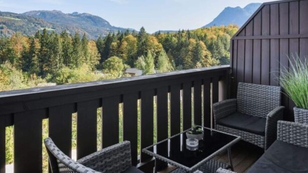 Ein Balkon mit Korbmöbeln, einem Glastisch und Blick auf einen Wald und Berge.