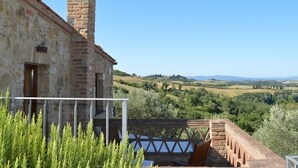 Un bâtiment en pierre doté d’une cheminée, d’un balcon avec une balustrade et offrant une vue sur des collines ondoyantes.