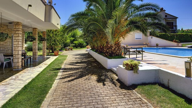 A poolside area with a paved walkway, palm trees, and outdoor furniture. - Apartment in Medulin Near the Beach (Medulin)