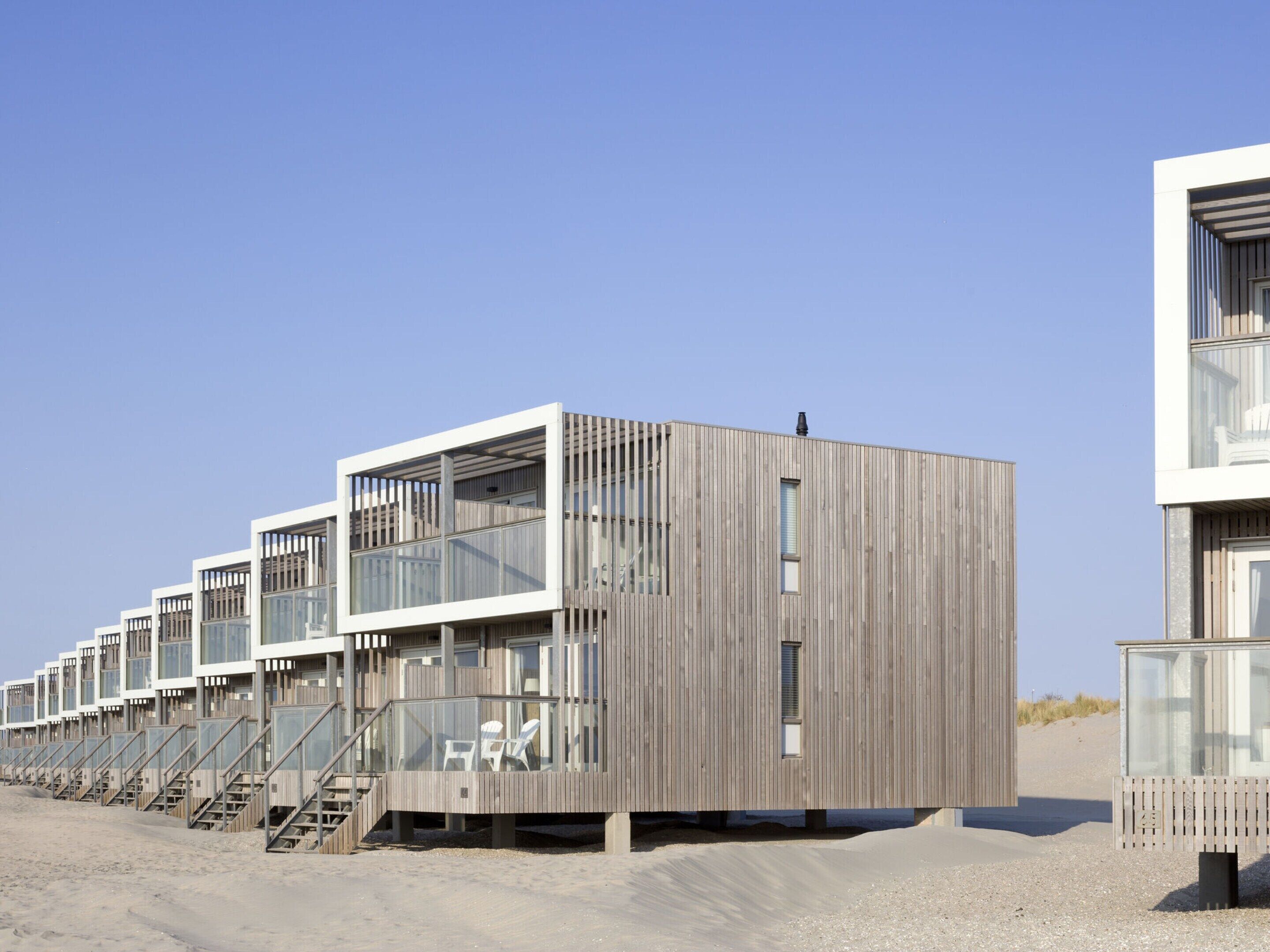 A row of wooden beach houses with glass balconies, steps leading to the beach, and a clear sky.