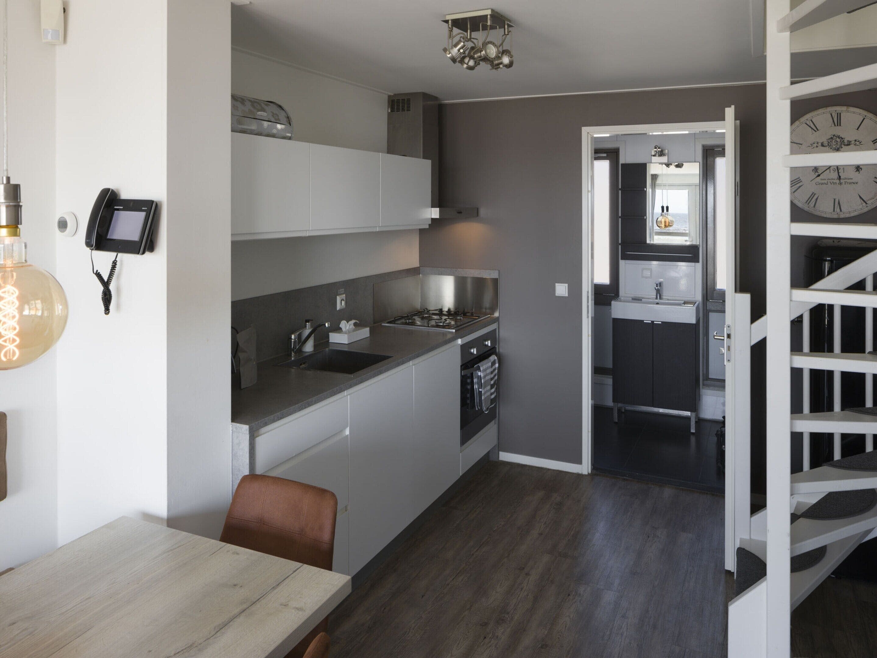 A modern kitchen with white cabinets, a built-in oven, and a sink. There is a dining table with chairs and a clock on the wall.