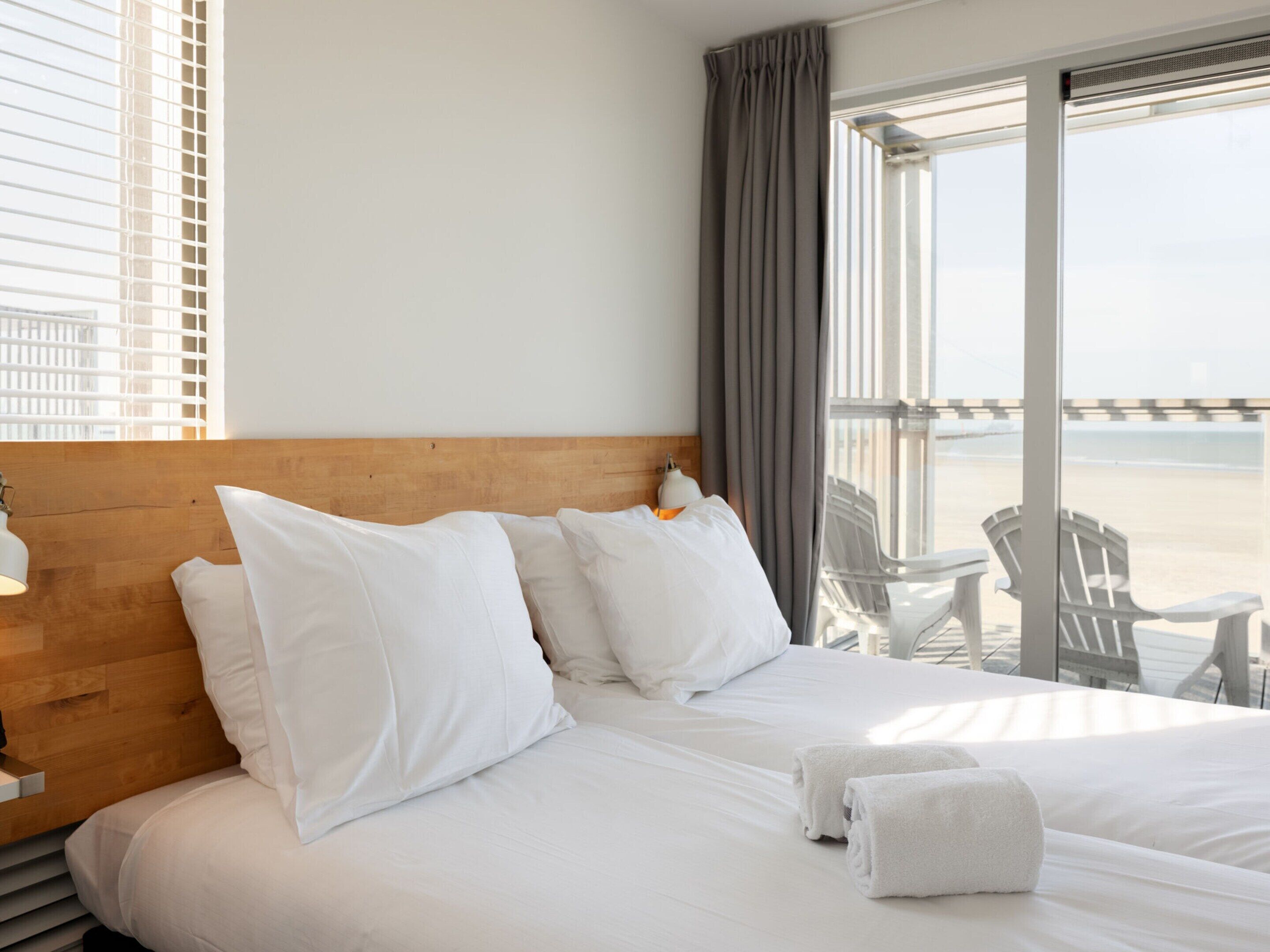 Une chambre d’hôtel avec un grand lit, du linge de lit blanc, une tête de lit en bois, une lampe de chevet et une vue sur la plage par une grande fenêtre.