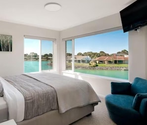 A bedroom with a large bed, a blue armchair, and a view of a lake and buildings.