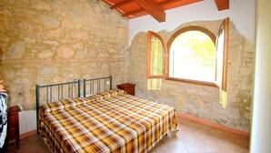 A bedroom with a stone wall, a wooden ceiling, a bed with a plaid bedspread, and a window with a view of greenery. - Belvilla by OYO Tuscan Farmhouse With Private Pool (Montescudaio)