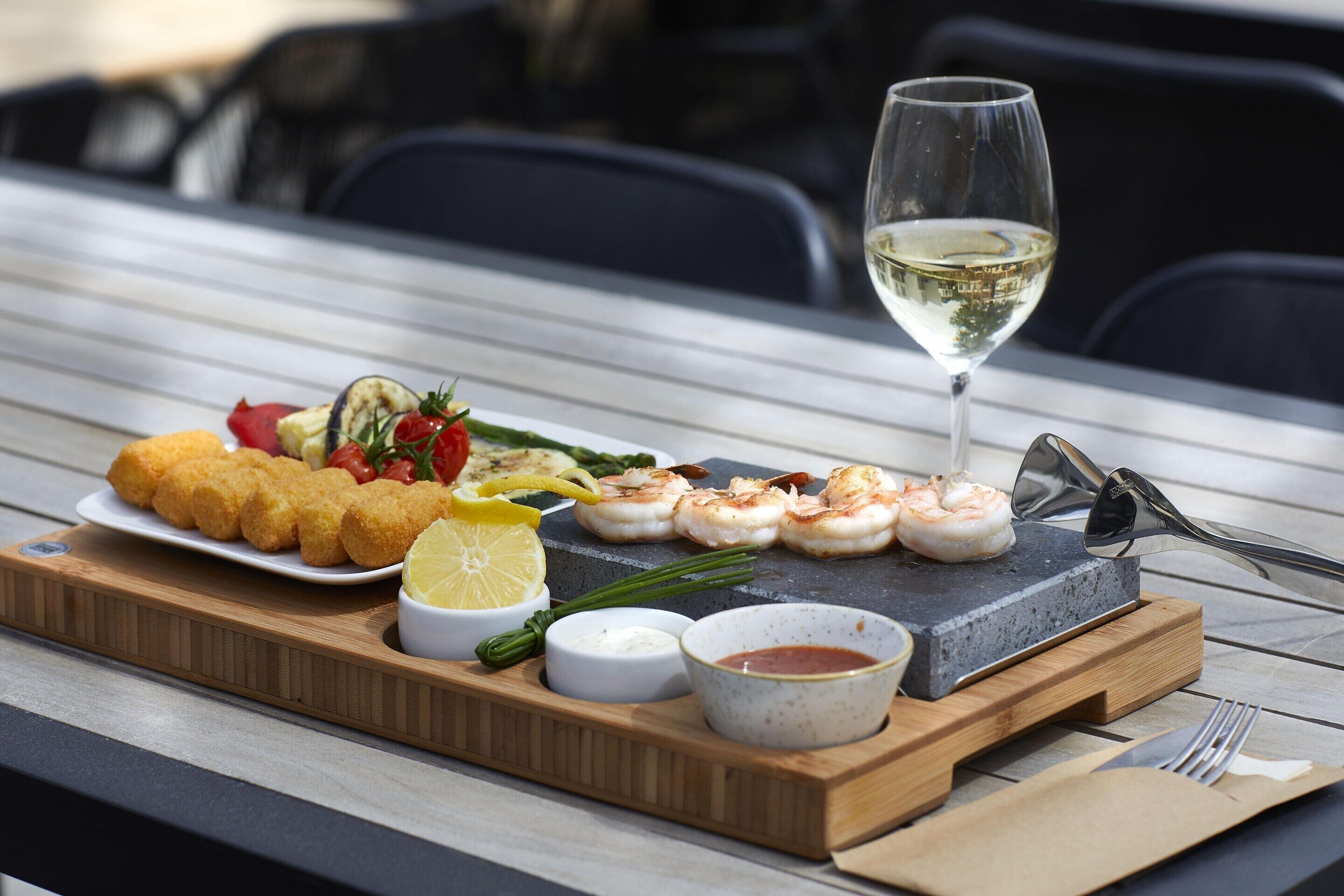 A meal with shrimp, vegetables, and bread on a wooden board, accompanied by a glass of white wine and condiments.