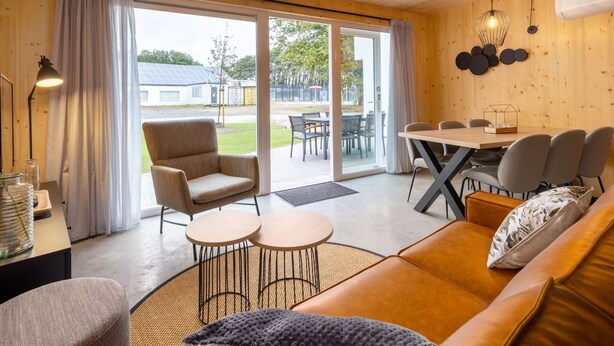 A cozy room with a sofa, a dining table, and a chair. There is a window with curtains, and a lamp is on a table.