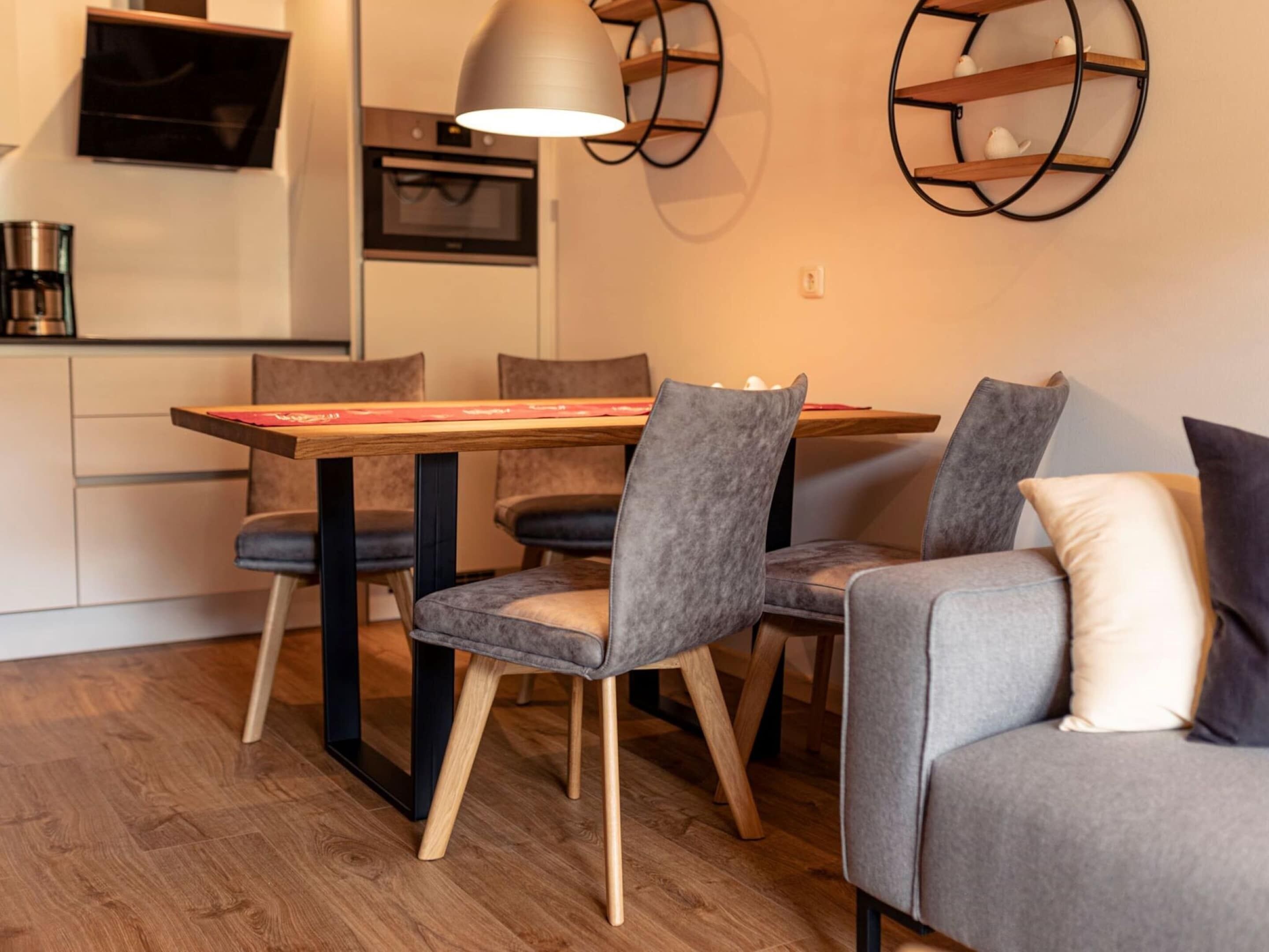 A modern kitchen with a dining area, featuring a wooden table, grey upholstered chairs, and a grey sofa with a white pillow.