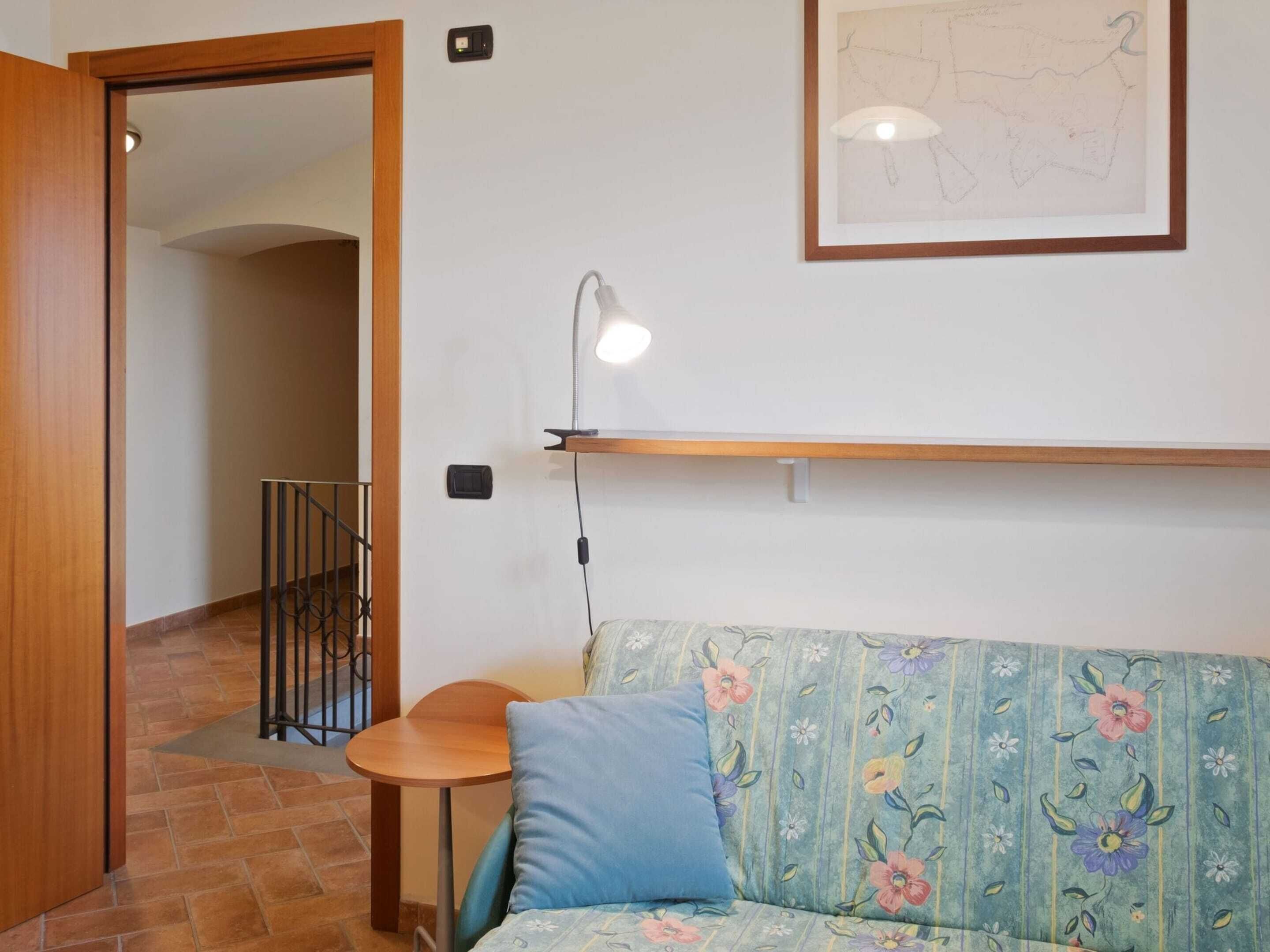 A room with a wooden door, a small round table, a sofa with floral upholstery, a wall-mounted shelf, and a framed map.