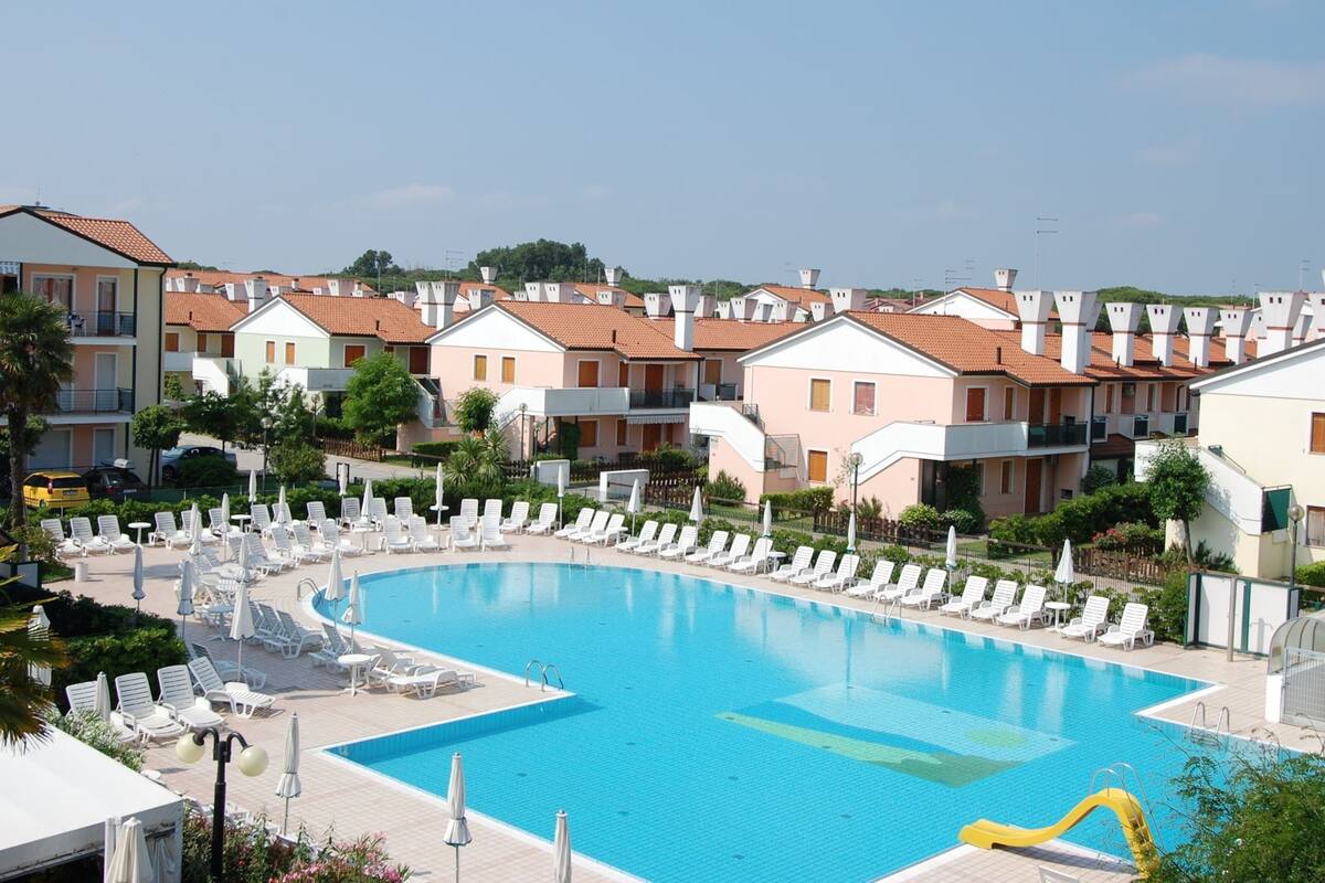 A swimming pool surrounded by white lounge chairs and umbrellas, with terraced buildings in the background.