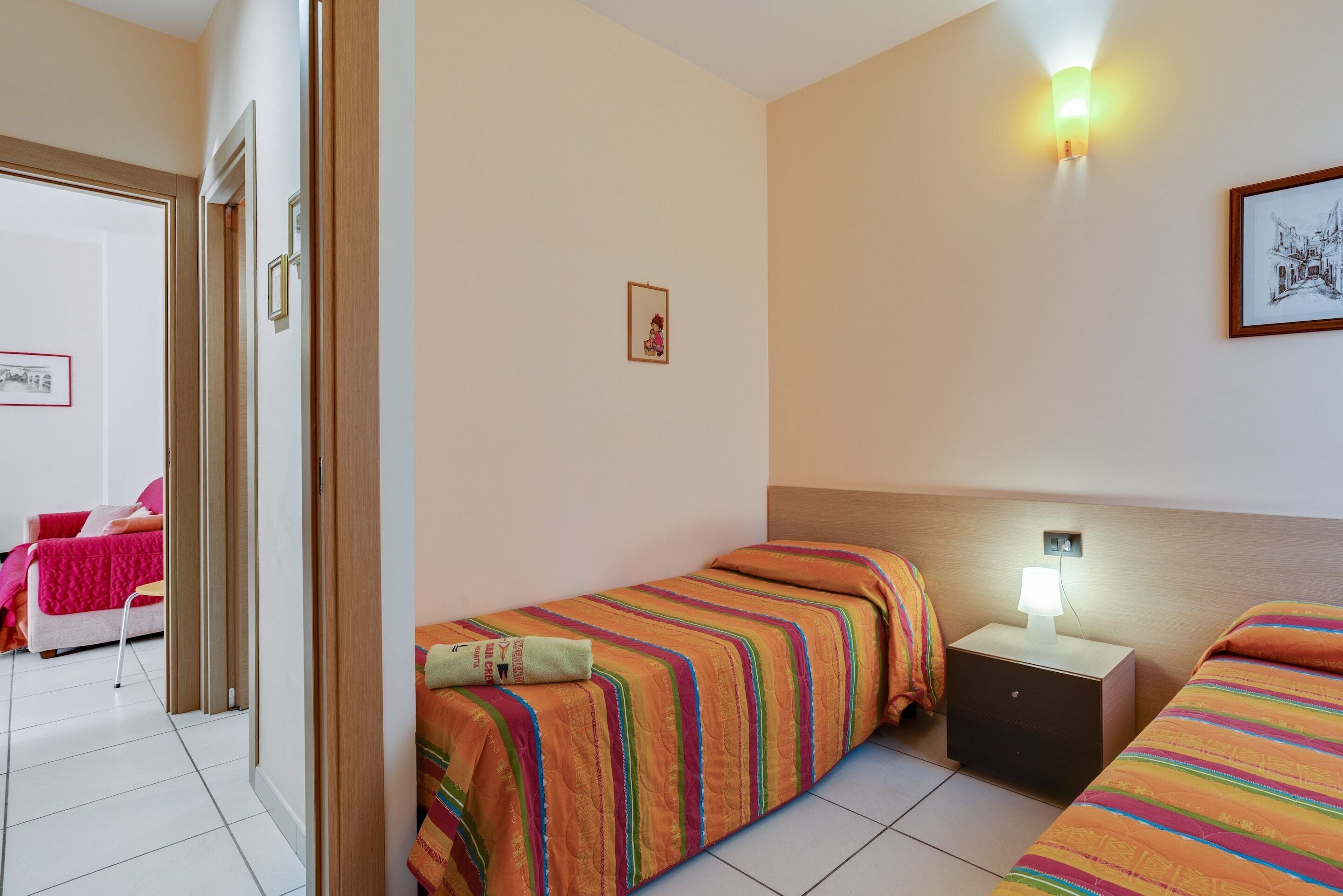 A hotel room with two single beds, a nightstand with a lamp, a door leading to another room, and a wall-mounted picture.