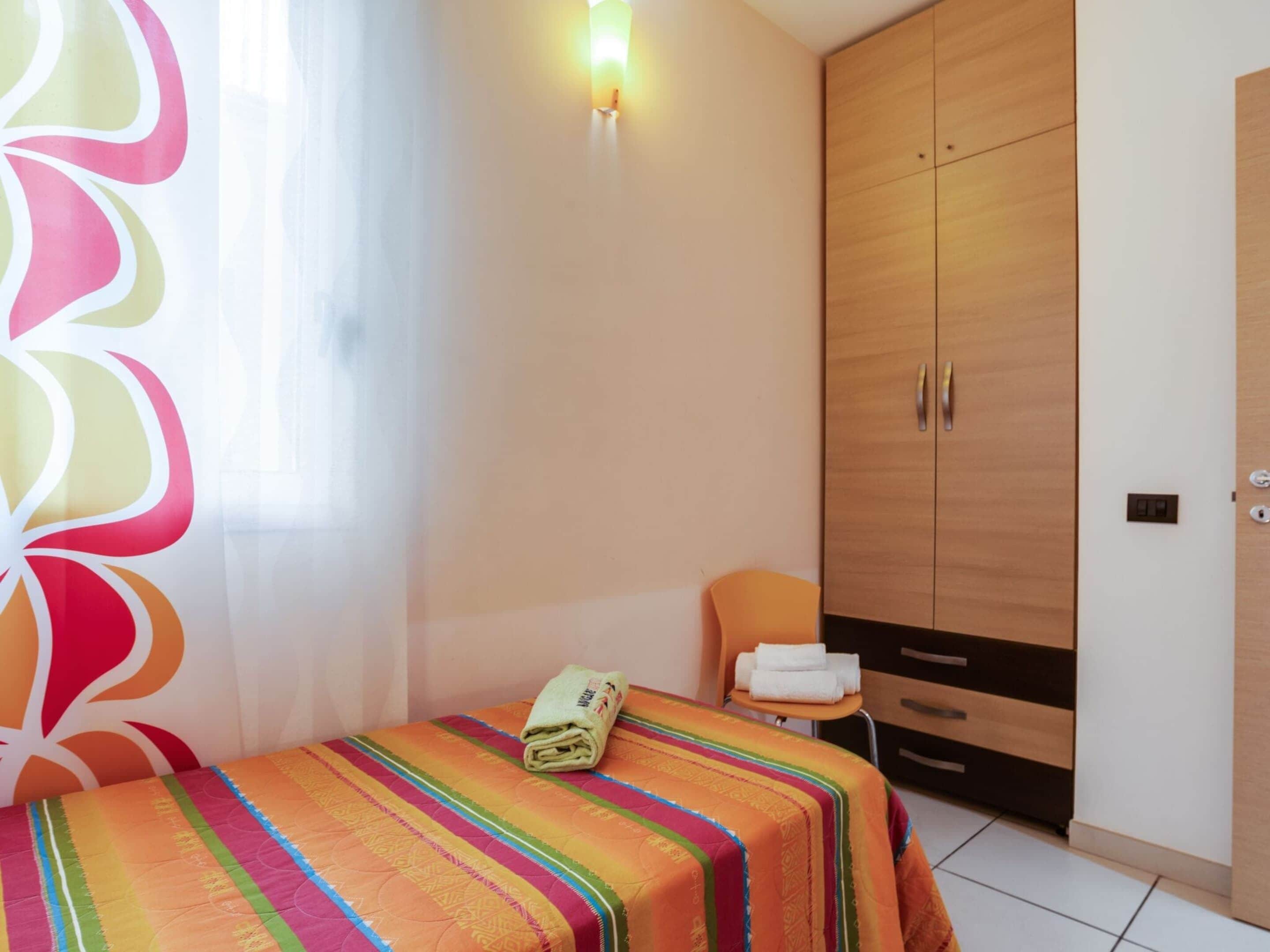 A bedroom with a bed, a wooden wardrobe, a chair, and a wall-mounted light.
