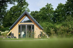 A modern, triangular wooden cabin with a glass door, surrounded by lush greenery.
