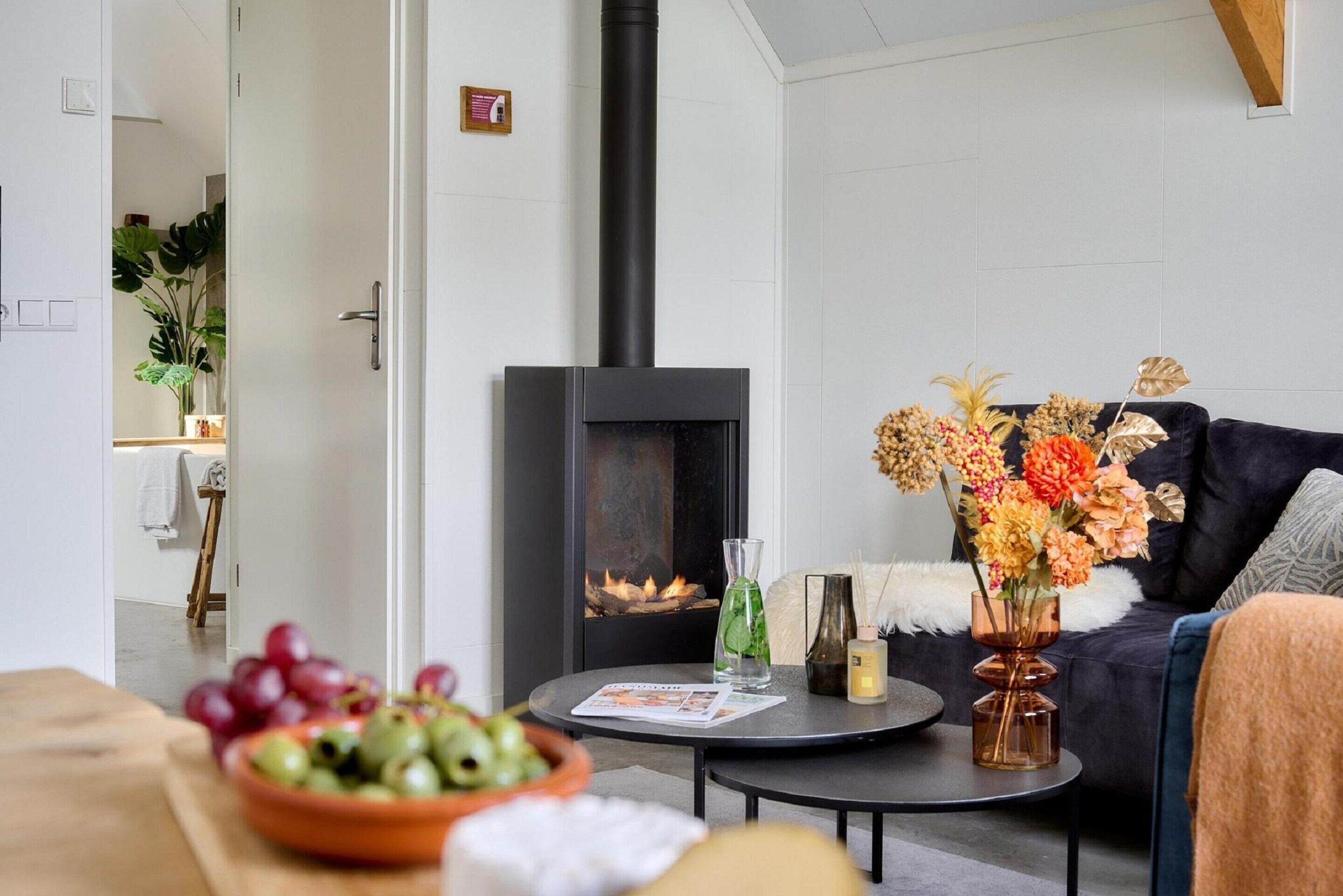 A modern living room with a black wood-burning stove, a dark sofa, a round coffee table with decorative items, and a bowl of fruit on a wooden table.