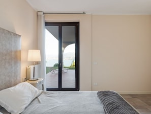 Habitación de hotel con cama, mesita de noche con lámpara, vista a un balcón con una silla y una puerta que da al exterior.