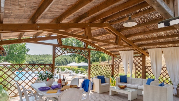 A covered outdoor seating area with wicker furniture, a table with food and drinks, and a view of a pool and greenery. - Belvilla by OYO Selvatello Villino Trilo (Casole d'Elsa)