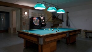 Une table de billard avec des billes, un porte-billes et deux jeux d’arcade dans une pièce dotée d’un escalier et d’un mur en pierre.