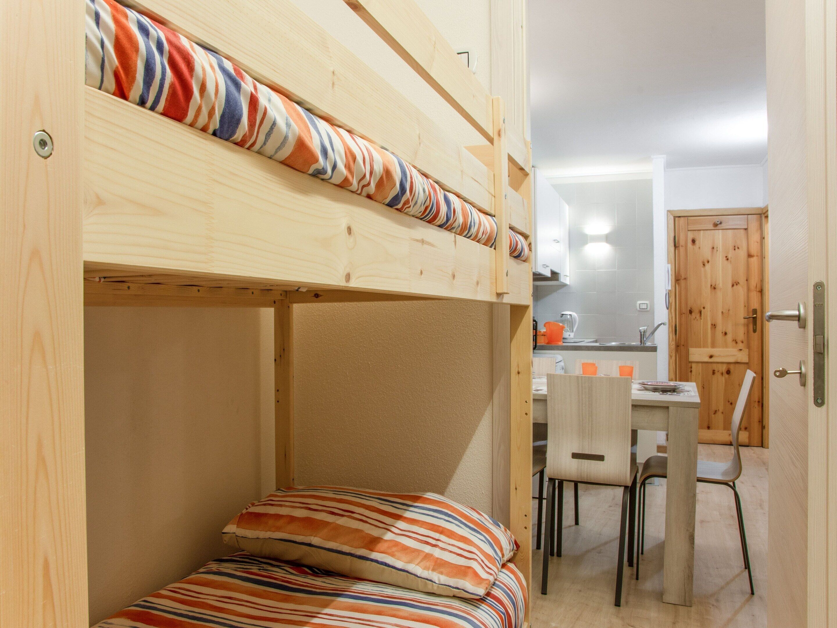 Uma cama beliche com roupa de cama listrada, uma escrivaninha de madeira com duas cadeiras e uma cozinha visível através de uma porta aberta.