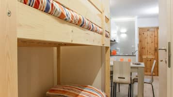 Uma cama beliche com roupa de cama listrada, uma escrivaninha de madeira com duas cadeiras e uma cozinha visível através de uma porta aberta.
