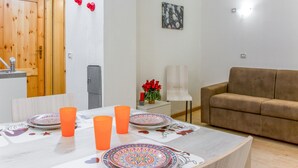 Un salon moderne avec un canapé marron, une table à manger ornée d’assiettes colorées et de tasses orange, une petite table d’appoint sur laquelle est posé un vase rempli de fleurs rouges, et une porte en bois.