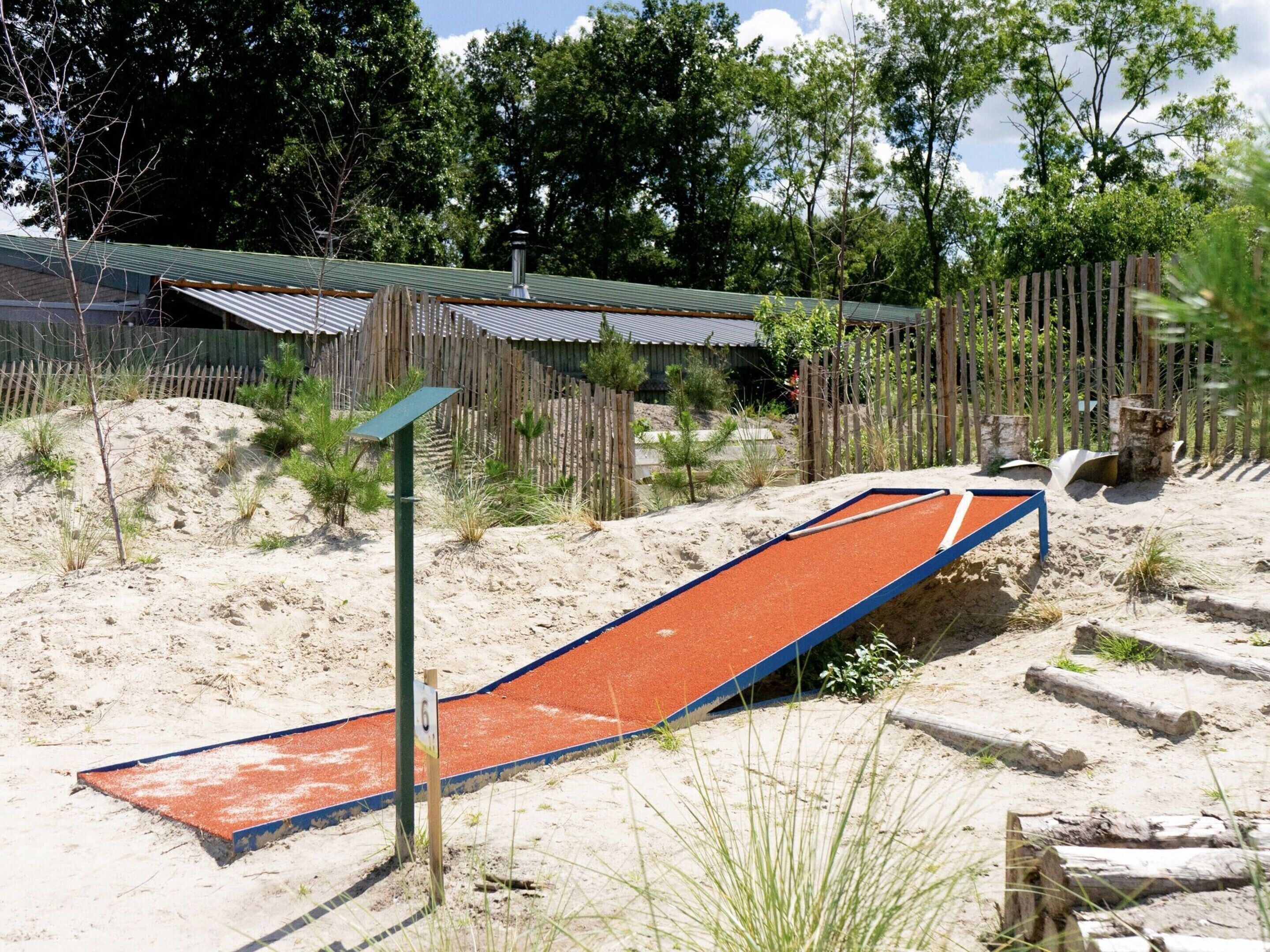 A sandy area with a small red slide, wooden fences, and a metal roof structure in the background.