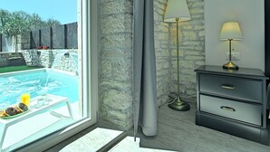 A room with a stone wall, a window with a view of a swimming pool, a bedside table with a lamp, and a dresser. - Perfect for Children, Garden With Pool, Playground and Walk in Whirlpool (Tinjan)