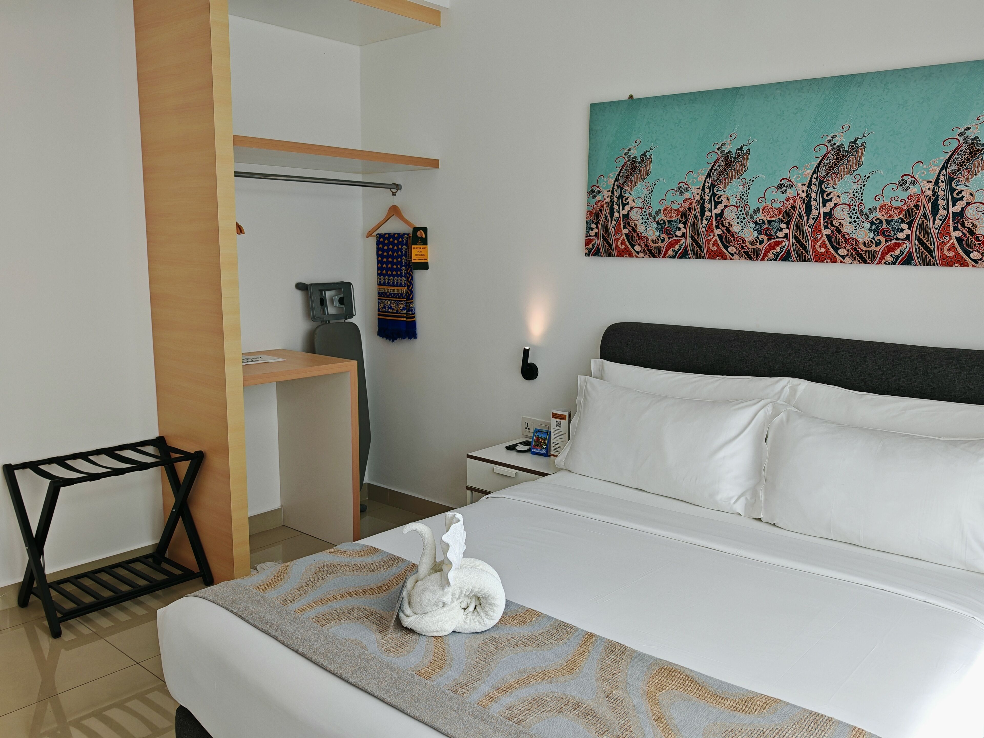 A hotel room with a bed, a desk, a chair, a coat rack, a painting, and a wall-mounted light.