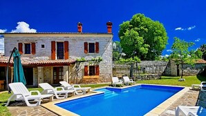 Un bâtiment en pierre doté d’une piscine, de chaises longues blanches et d’un parasol vert.