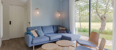 Een moderne woonkamer met een blauwe bank, twee stoelen, een klein houten tafeltje en een groot raam met uitzicht op groen.