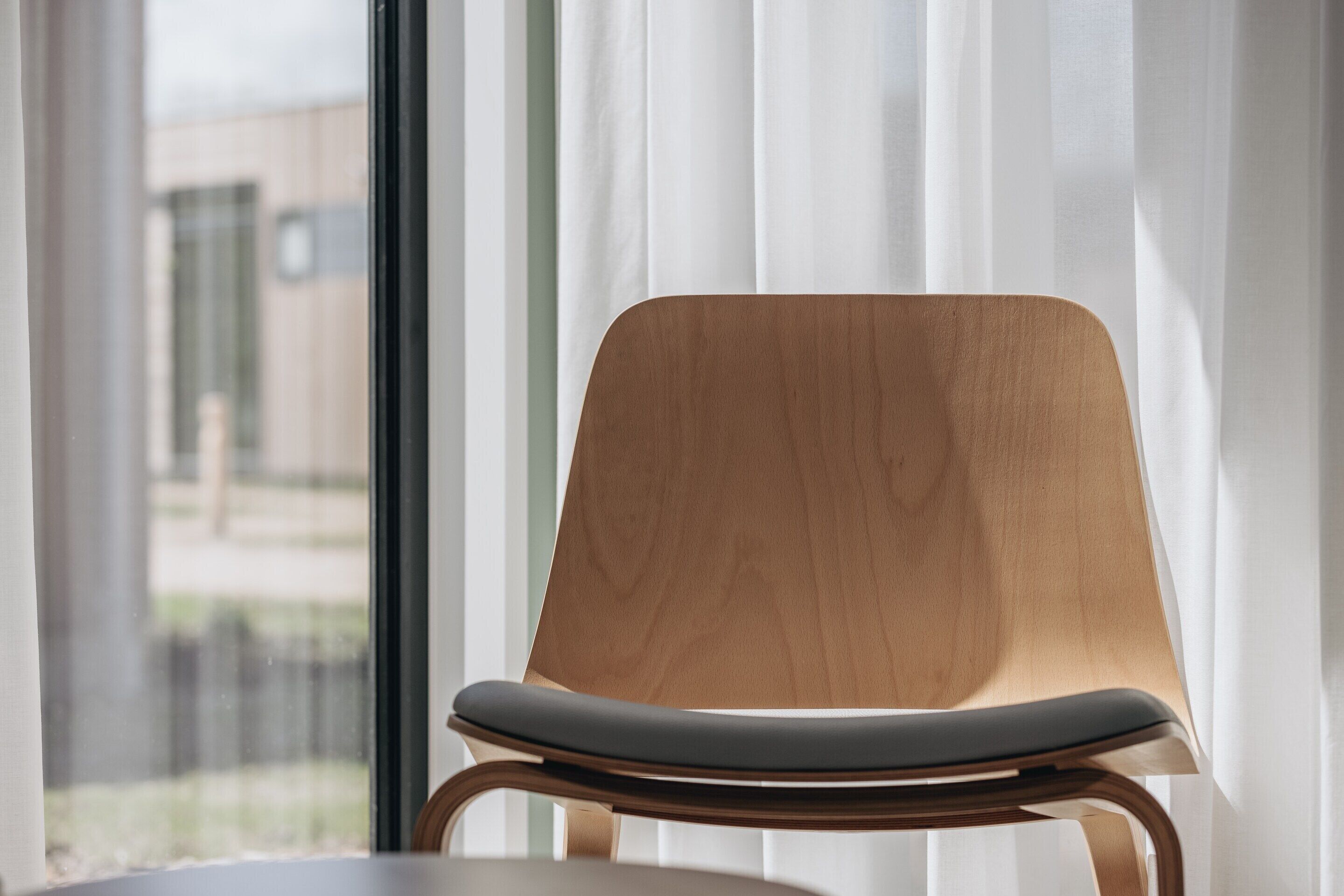 A modern chair with a wooden backrest and a cushioned seat, positioned near a window with sheer curtains.