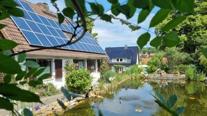 Een huis met zonnepanelen op het dak, een vijver en omliggend groen.