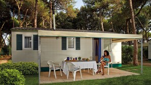 A small, single-story building with a covered patio, a table with chairs, and a person seated. - Cozy Pine Forest Chalet (Cellole)