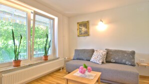 Ein gemütliches Wohnzimmer mit einer grauen Couch, zwei Topfpflanzen, einem kleinen Holztisch mit Blumen und einem an der Wand befestigten Bilderrahmen.