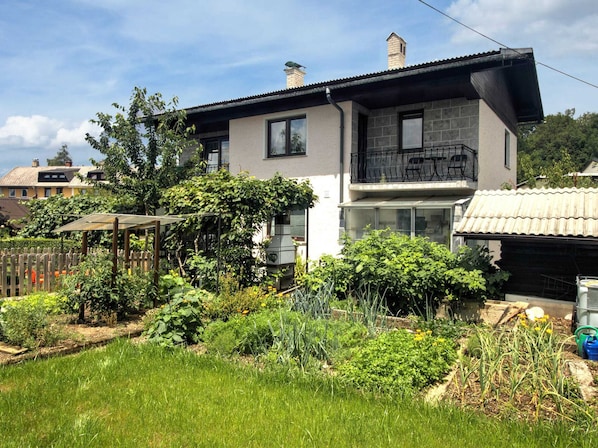 Una casa de dos pisos con un balcón, rodeada de un jardín con diversas plantas y una cerca de madera.