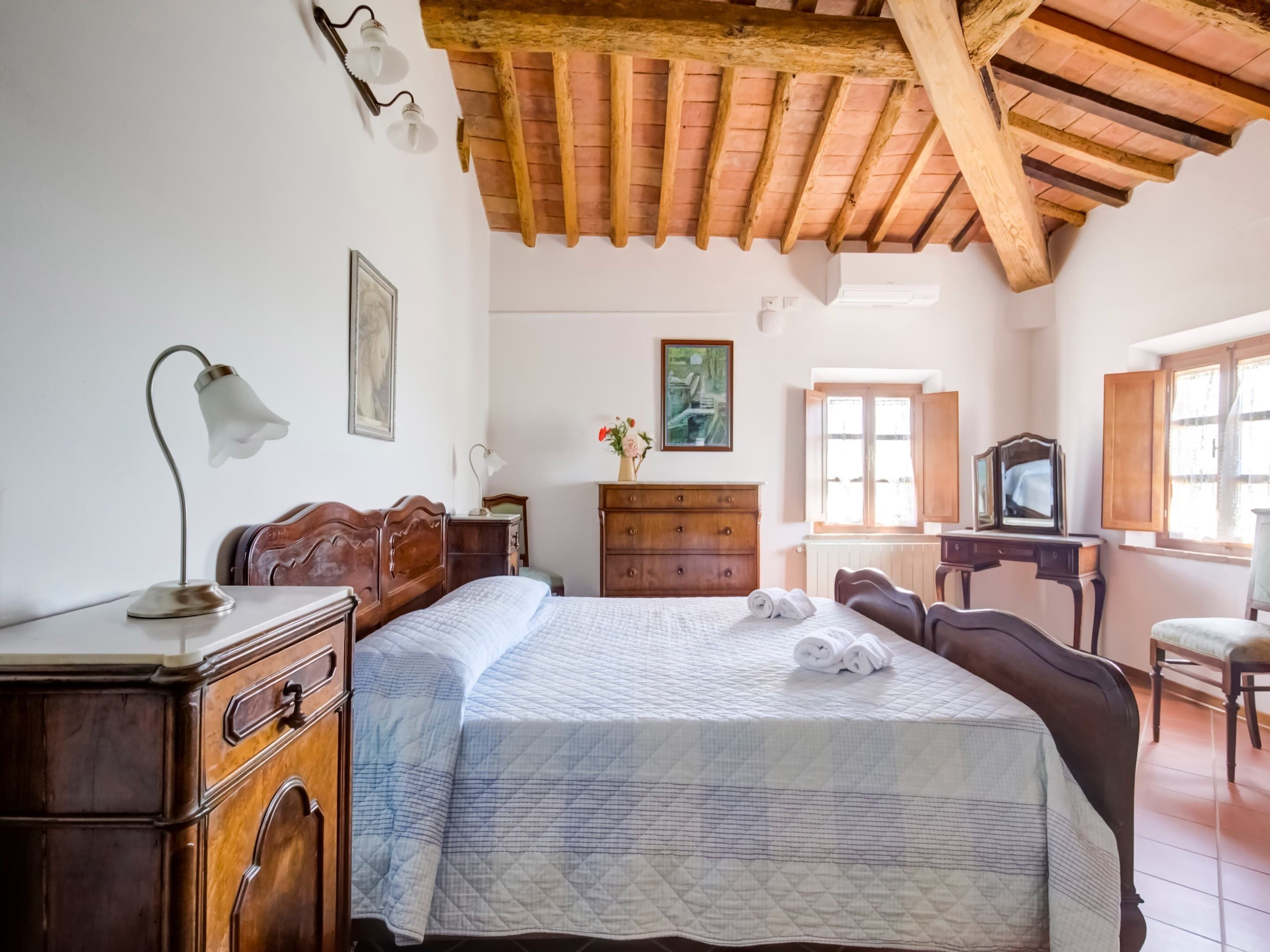 A bedroom with a wooden ceiling, a bed with a quilted bedspread, a dresser, a desk with a mirror, and a window with wooden shutters.
