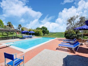Piscina con sillas reclinables azules y sombrillas, rodeada de follaje y un cielo despejado.