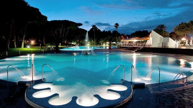 Une piscine nocturne avec une fontaine centrale, entourée d’une terrasse dallée et d’un bâtiment blanc en arrière-plan.