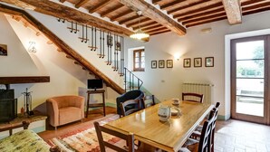 A spacious living area with a wooden dining table, comfortable seating, a staircase with exposed beams, and a view through a large window. - Belvilla by OYO Piandigoro - I Lecci (Radicofani)