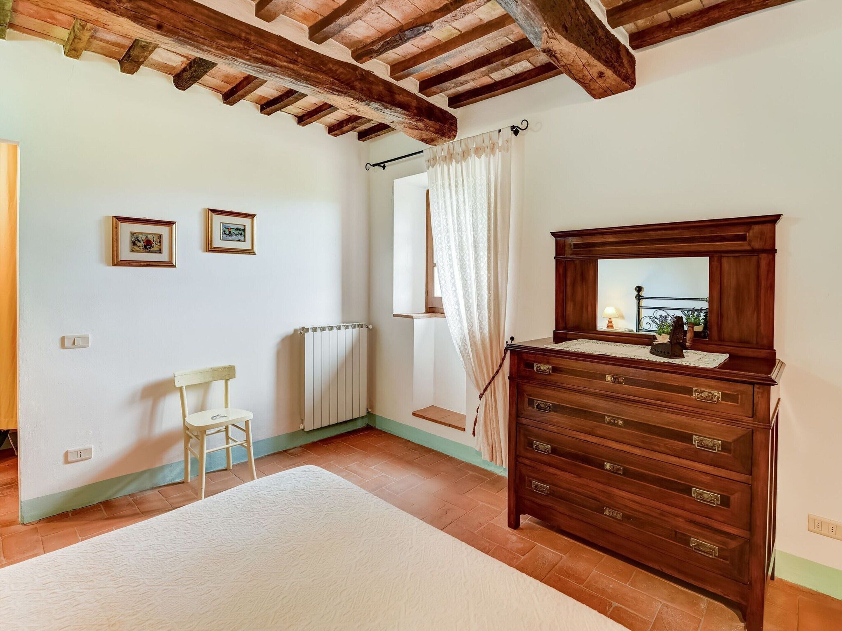 Un dormitorio con un aparador de madera, una silla, un radiador y una ventana con cortinas.