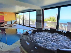 Uma banheira de hidromassagem em um quarto com janelas amplas, piscina e espreguiçadeiras.