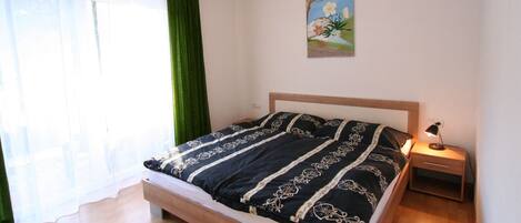 Un dormitorio con cama, mesita de noche, lámpara, un cuadro y una ventana con cortinas.