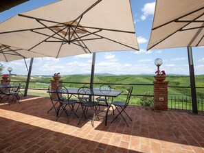 A patio with tables and chairs under large umbrellas, overlooking a green landscape with hills.