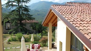 Une terrasse avec des tables et des chaises rouges, un bâtiment jaune avec un toit en tuiles, et un paysage montagneux en arrière-plan.